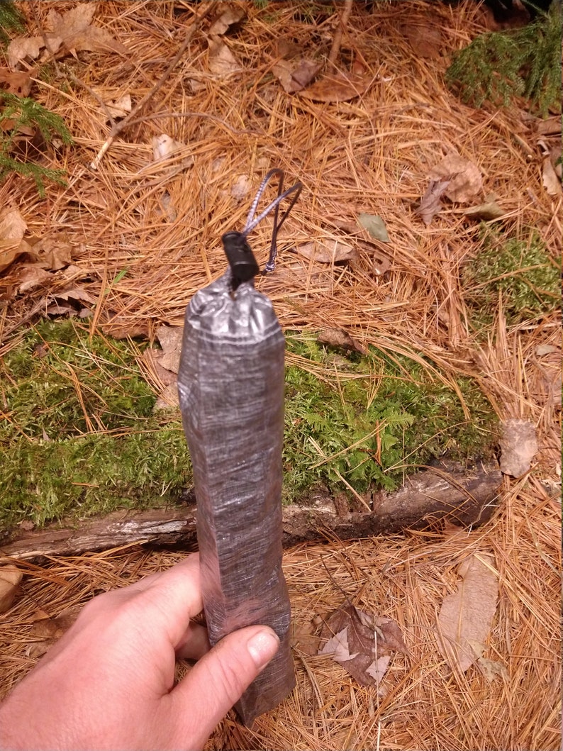 Puede incluir: Un objeto rectangular gris oscuro envuelto en cinta adhesiva, con un cierre de cord&oacute;n negro. El objeto se sostiene sobre un fondo de agujas de pino, hojas y musgo.