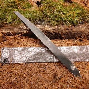 Puede incluir: Una sierra met&aacute;lica con una hoja dentada descansa sobre una funda plateada, sobre un fondo de agujas de pino y un tronco cubierto de musgo. La sierra parece usada, lo que sugiere aplicaciones al aire libre o de carpinter&iacute;a.