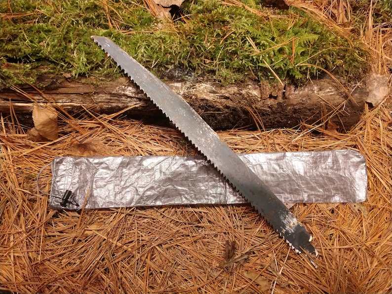 Puede incluir: Una sierra met&aacute;lica con una hoja dentada descansa sobre una funda plateada, sobre un fondo de agujas de pino y un tronco cubierto de musgo. La sierra parece usada, lo que sugiere aplicaciones al aire libre o de carpinter&iacute;a.