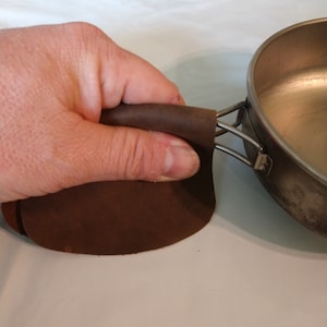 May include: A brown leather handle for a camping pot or pan. The handle is wrapped around the metal handle of the pot.