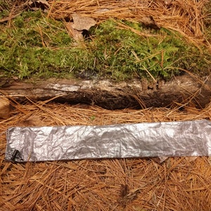 Puede incluir: Una manta de supervivencia plateada y rectangular est&aacute; colocada sobre un lecho de agujas de pino. Un tronco cubierto de musgo verde est&aacute; en el fondo. La manta tiene un cable y un conector negros.