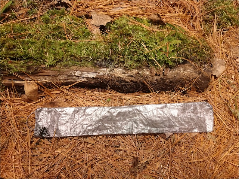 Puede incluir: Una manta de supervivencia plateada y rectangular est&aacute; colocada sobre un lecho de agujas de pino. Un tronco cubierto de musgo verde est&aacute; en el fondo. La manta tiene un cable y un conector negros.