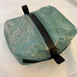 May include: A small, teal-colored zippered pouch with a black zipper and black straps. The pouch is made of a lightweight, water-resistant material.