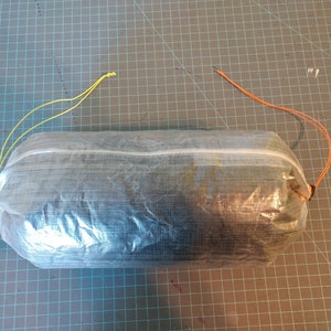 May include: A clear, lightweight, and compact stuff sack with a white zipper and two drawstrings, one yellow and one orange, for securing the contents.