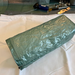 May include: A rectangular, sage green, crinkled fabric item, possibly a bag or a wrapped item, rests on a white fabric surface. The item has a shiny, textured appearance. Sewing tools and materials are visible in the background.