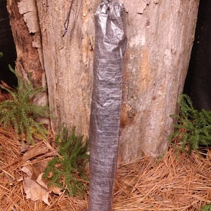 Puede incluir: Una bolsa cil&iacute;ndrica gris oscuro con cierre de cord&oacute;n. La bolsa est&aacute; hecha de un material ligero y arrugado. El fondo presenta un tronco de &aacute;rbol y suelo forestal.