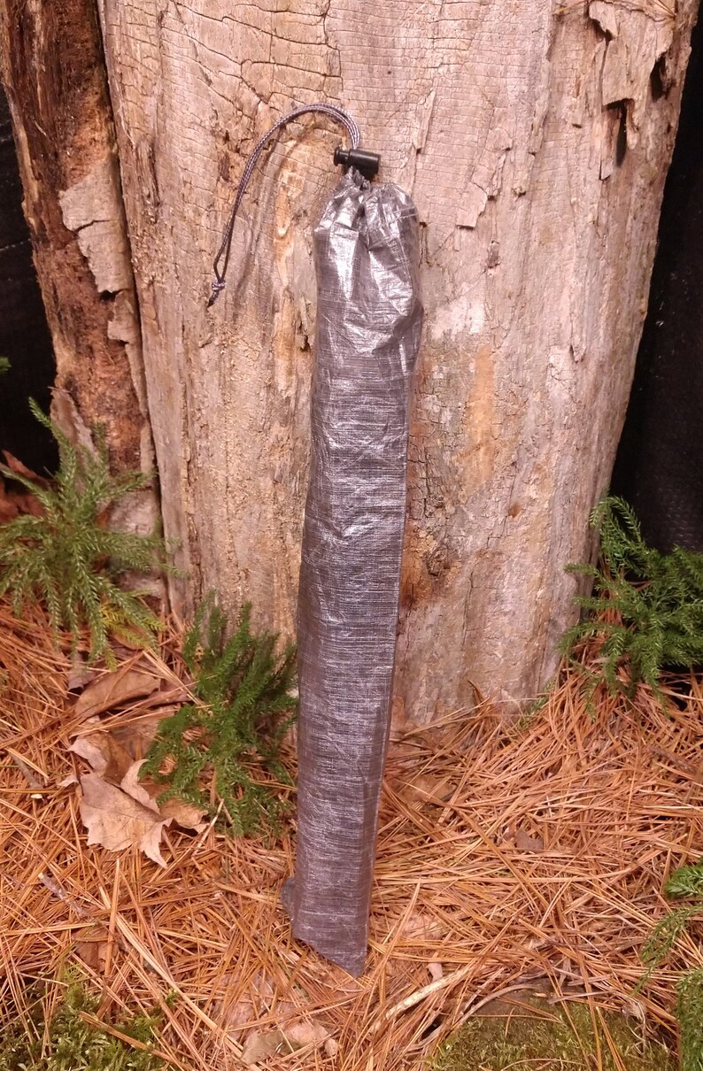 Puede incluir: Una bolsa cil&iacute;ndrica gris oscuro con cierre de cord&oacute;n. La bolsa est&aacute; hecha de un material ligero y arrugado. El fondo presenta un tronco de &aacute;rbol y suelo forestal.