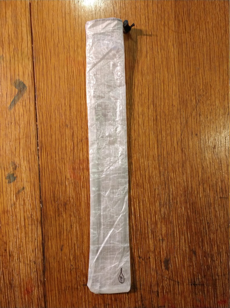 Puede incluir: Una bolsa rectangular transl&uacute;cida con cierre de cord&oacute;n. La bolsa est&aacute; hecha de un material ligero y arrugado. Un peque&ntilde;o gr&aacute;fico negro est&aacute; impreso cerca de la parte inferior. La bolsa est&aacute; sobre una superficie de madera.