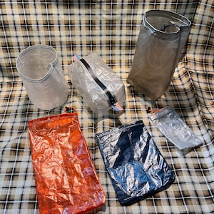 May include: Six lightweight, translucent bags made of ripstop nylon in various colours, including orange, blue, and grey. The bags are all folded and laid out on a plaid fabric background.