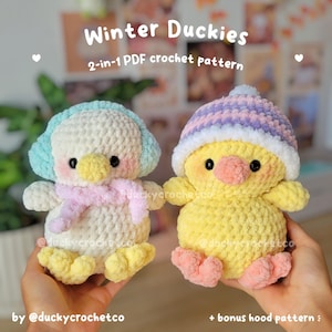 May include: Two plush crochet duckies. The left duck is white with light blue earmuffs and a pink scarf. The right duck is yellow with a purple and white striped hat. Text reads: Winter Duckies 2-in-1 PDF crochet pattern + bonus hood pattern.