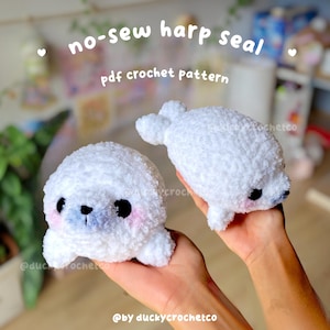 May include: Two white plush harp seal toys made of soft, fuzzy yarn. Each seal features large black safety eyes, a small black nose, and pink blush on the cheeks. The text "no-sew harp seal pdf crochet pattern" and "@by duckycrochetco" is visible.
