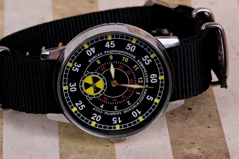 May include: A black wristwatch with a silver case and a black nylon strap. The watch face has a black background with white and yellow markings. The watch face also has a small yellow and black radiation symbol. The watch face has the text "ВОЙСКА РАДИАЦИОННОЙ, ХИМИЧЕСКОЙ И БИОЛОГИЧЕСКОЙ ЗАЩИТЫ СССР" in Cyrillic.