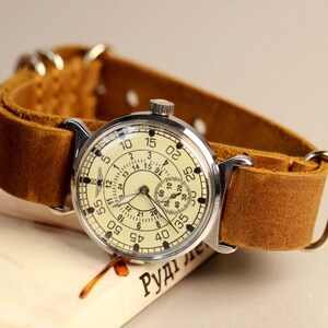 Puede incluir: Un reloj de pulsera vintage con una caja plateada y una esfera de color crema. El reloj tiene una correa de cuero marr&oacute;n y una esfera de 24 horas. La esfera del reloj tiene n&uacute;meros ar&aacute;bigos negros y una aguja de segundos negra.