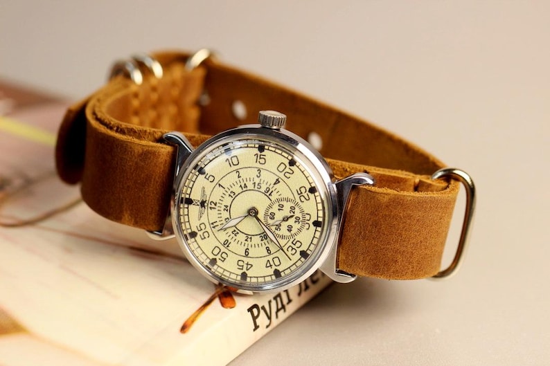 Puede incluir: Un reloj de pulsera vintage con una caja plateada y una esfera de color crema. El reloj tiene una correa de cuero marr&oacute;n y una esfera de 24 horas. La esfera del reloj tiene n&uacute;meros ar&aacute;bigos negros y una aguja de segundos negra.