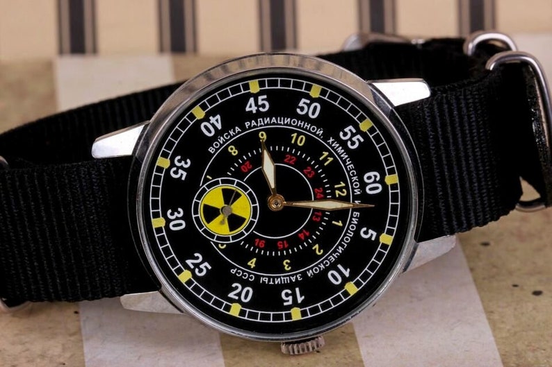 May include: A black wristwatch with a silver case and a black nylon strap. The watch face has a black background with white and yellow markings. The watch face also has a smaller circular dial with a yellow and black radiation symbol. The watch face has the text "ВОЙСКА РАДИАЦИОННОЙ, ХИМИЧЕСКОЙ И БИОЛОГИЧЕСКОЙ ЗАЩИТЫ СССР" around the outer edge.