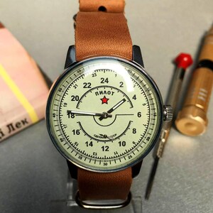 Puede incluir: Un reloj de pulsera vintage con una correa de cuero marr&oacute;n. La esfera del reloj es blanca con marcas negras y una estrella roja. La esfera del reloj dice "Пилот", que en ruso significa "Piloto".