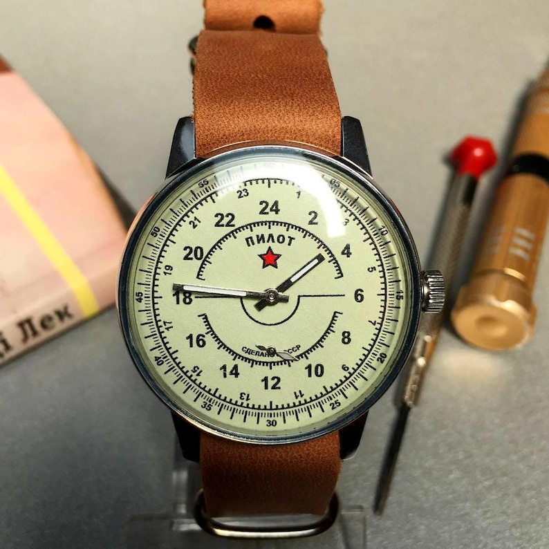 Puede incluir: Un reloj de pulsera vintage con una correa de cuero marr&oacute;n. La esfera del reloj es blanca con marcas negras y una estrella roja. La esfera del reloj dice "Пилот", que en ruso significa "Piloto".