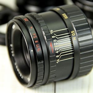 Puede incluir: Un objetivo de c&aacute;mara negro con marcas rojas y blancas, incluyendo n&uacute;meros de diafragma y escalas de distancia. El objetivo tiene un anillo de enfoque estriado y una escala de profundidad de campo. Otros objetivos negros est&aacute;n en el fondo.