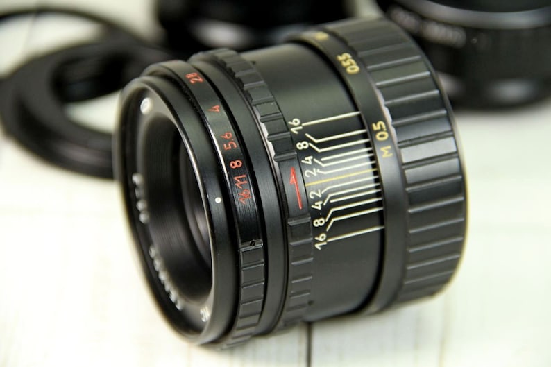 Puede incluir: Un objetivo de c&aacute;mara negro con marcas rojas y blancas, incluyendo n&uacute;meros de diafragma y escalas de distancia. El objetivo tiene un anillo de enfoque estriado y una escala de profundidad de campo. Otros objetivos negros est&aacute;n en el fondo.