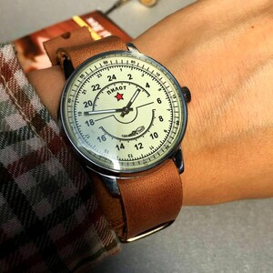 Puede incluir: Un reloj de pulsera con una correa de cuero marr&oacute;n y una esfera blanca. La esfera tiene marcas negras y una estrella roja con la palabra "ПИЛОТ" escrita en cir&iacute;lico. El reloj est&aacute; en la mu&ntilde;eca de una persona.