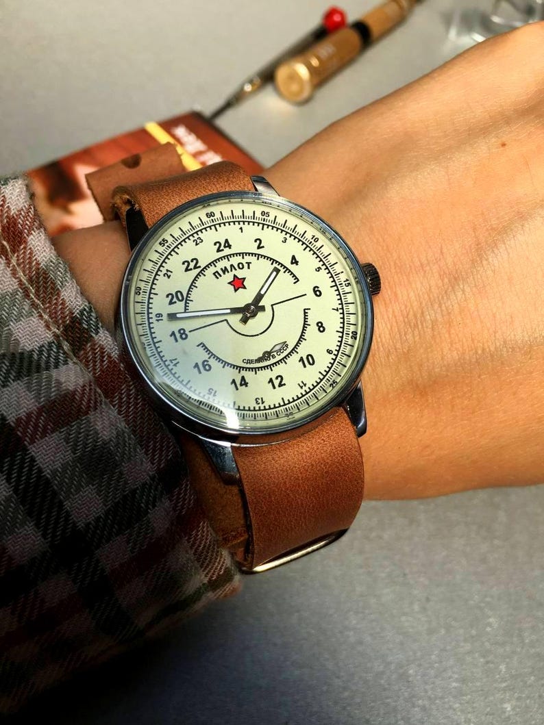 Puede incluir: Un reloj de pulsera con una correa de cuero marr&oacute;n y una esfera blanca. La esfera tiene marcas negras y una estrella roja con la palabra "ПИЛОТ" escrita en cir&iacute;lico. El reloj est&aacute; en la mu&ntilde;eca de una persona.