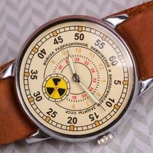 May include: A vintage wristwatch with a white face and black hands. The watch face has a yellow radiation symbol and the text "ВОЙСКА РАДИАЦИОННОЙ, ХИМИЧЕСКОЙ И БИОЛОГИЧЕСКОЙ ЗАЩИТЫ СССР" in Cyrillic script.