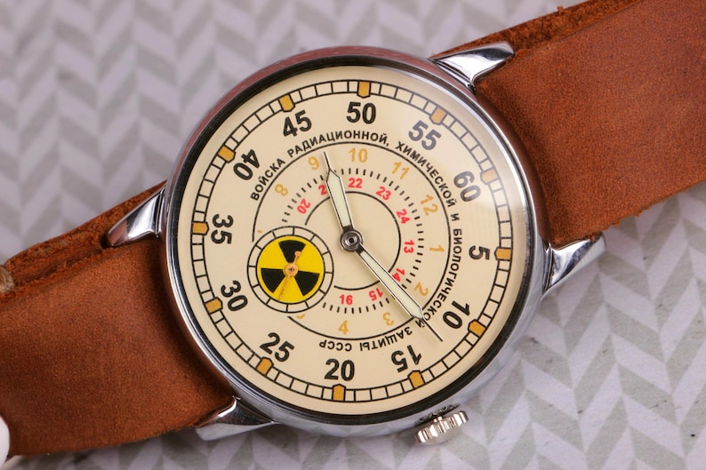 May include: A vintage wristwatch with a white face and black hands. The watch face has a yellow radiation symbol and the text "ВОЙСКА РАДИАЦИОННОЙ, ХИМИЧЕСКОЙ И БИОЛОГИЧЕСКОЙ ЗАЩИТЫ СССР" in Cyrillic script.