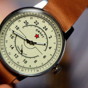 Puede incluir: Un reloj de pulsera anal&oacute;gico blanco con una correa de cuero marr&oacute;n. La esfera del reloj tiene marcas negras y una estrella roja con la palabra "ПИЛОТ" en escritura cir&iacute;lica. La esfera del reloj tambi&eacute;n tiene la palabra "СССР" en escritura cir&iacute;lica.
