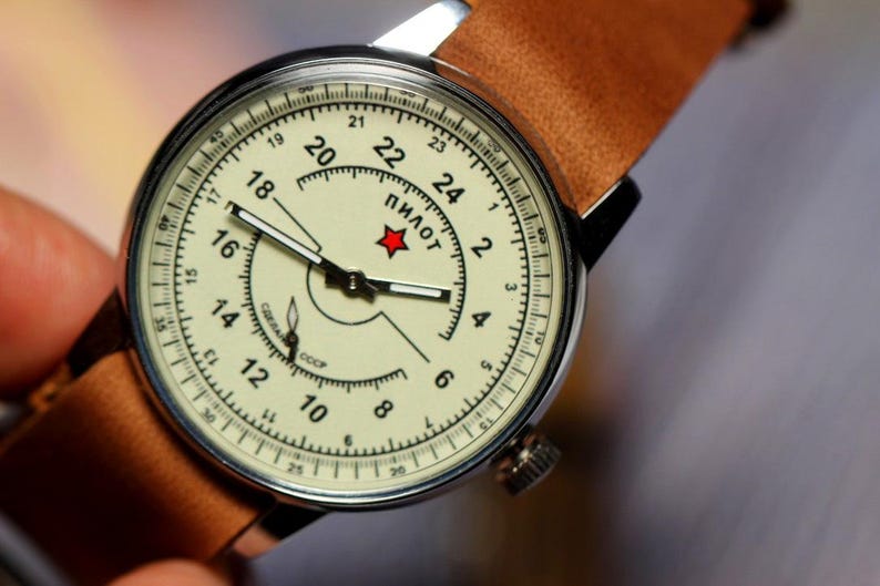 Puede incluir: Un reloj de pulsera anal&oacute;gico blanco con una correa de cuero marr&oacute;n. La esfera del reloj tiene marcas negras y una estrella roja con la palabra "ПИЛОТ" en escritura cir&iacute;lica. La esfera del reloj tambi&eacute;n tiene la palabra "СССР" en escritura cir&iacute;lica.