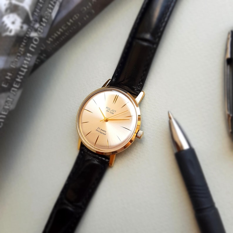 K&ouml;nnte beinhalten: Eine goldfarbene Armbanduhr mit einem schwarzen Lederarmband. Das Zifferblatt der Uhr ist wei&szlig; mit schwarzen Markierungen und dem aufgedruckten Text "Poljot 22 Jewels Shockproof".
