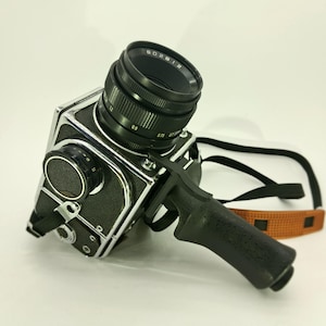 May include: A medium format film camera with a black textured body and chrome accents. It features a large lens with visible focus markings, a side-mounted black pistol grip handle, and a black neck strap with a tan textured section attached to the side.