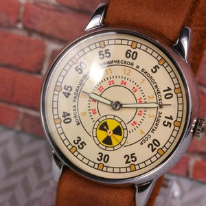 May include: A vintage wristwatch with a white face and black hands. The watch face has a red and black radiation symbol and the text "ВОЙСКА РАДИАЦИОННОЙ И БИОЛОГИЧЕСКОЙ ЗАЩИТЫ СССР" around the outer edge.