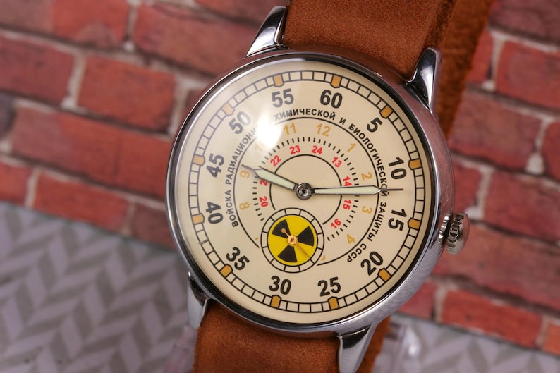 May include: A vintage wristwatch with a white face and black hands. The watch face has a red and black radiation symbol and the text "ВОЙСКА РАДИАЦИОННОЙ И БИОЛОГИЧЕСКОЙ ЗАЩИТЫ СССР" around the outer edge.