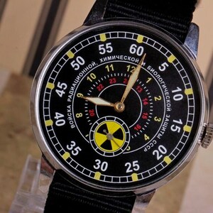 May include: A black wristwatch with a silver case and a black nylon strap. The watch face has a black background with yellow and red markings. The watch face also has a yellow and black radiation symbol. The text on the watch face reads "Войска радиационной, химической и биологической защиты СССР".