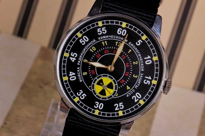 May include: A black wristwatch with a silver case and a black nylon strap. The watch face has a black background with yellow and red markings. The watch face also has a yellow and black radiation symbol. The text on the watch face reads "Войска радиационной, химической и биологической защиты СССР".
