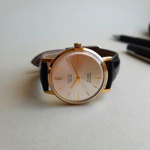K&ouml;nnte beinhalten: Eine goldfarbene Armbanduhr mit wei&szlig;em Zifferblatt und schwarzem Lederarmband. Das Zifferblatt der Uhr hat goldfarbene Zeiger und Markierungen sowie den aufgedruckten Text "Poljot Deluxe 23 Jewels Shockproof".