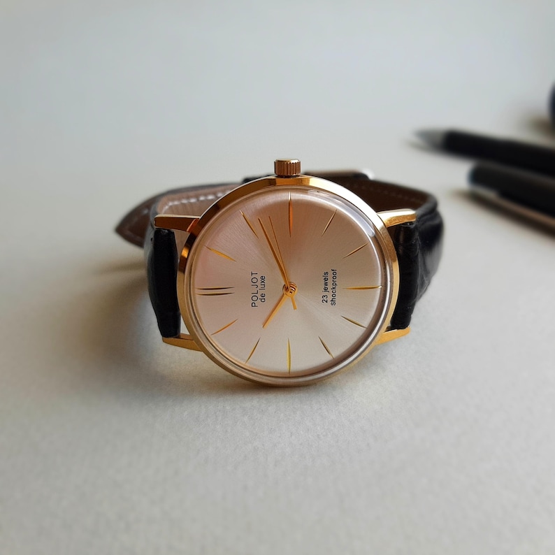 K&ouml;nnte beinhalten: Eine goldfarbene Armbanduhr mit wei&szlig;em Zifferblatt und schwarzem Lederarmband. Das Zifferblatt der Uhr hat goldfarbene Zeiger und Markierungen sowie den aufgedruckten Text "Poljot Deluxe 23 Jewels Shockproof".