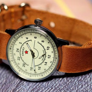 Puede incluir: Un reloj de pulsera vintage con una esfera blanca y agujas negras. El reloj tiene una correa de cuero marr&oacute;n y una caja plateada. La esfera del reloj tiene n&uacute;meros negros y la palabra "пилот" impresa en escritura cir&iacute;lica.