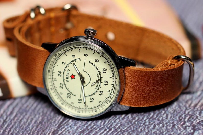 Puede incluir: Un reloj de pulsera vintage con una esfera blanca y agujas negras. El reloj tiene una correa de cuero marr&oacute;n y una caja plateada. La esfera del reloj tiene n&uacute;meros negros y la palabra "пилот" impresa en escritura cir&iacute;lica.
