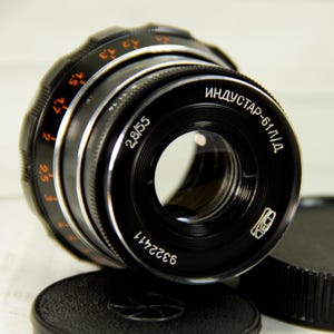 May include: A black Industar-61 L/D 2.8/55 camera lens with a black lens cap. The lens has white and orange markings, including the text "ИНДУСТАР-61 Л/Д" and the serial number 9322411. The lens is designed for photography.