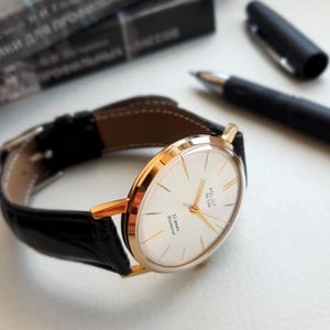 K&ouml;nnte beinhalten: Eine goldfarbene Armbanduhr mit wei&szlig;em Zifferblatt und schwarzem Lederarmband. Das Zifferblatt der Uhr hat schwarze r&ouml;mische Ziffern als Stundenmarkierungen und einen goldfarbenen Sekundenzeiger. Das Zifferblatt der Uhr tr&auml;gt au&szlig;erdem den Text "Poljot 26 Jewels" und "23 Jewels Shockproof".