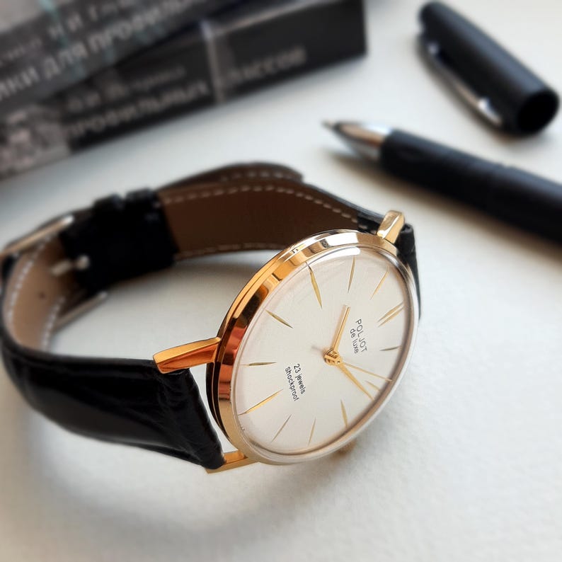 K&ouml;nnte beinhalten: Eine goldfarbene Armbanduhr mit wei&szlig;em Zifferblatt und schwarzem Lederarmband. Das Zifferblatt der Uhr hat schwarze r&ouml;mische Ziffern als Stundenmarkierungen und einen goldfarbenen Sekundenzeiger. Das Zifferblatt der Uhr tr&auml;gt au&szlig;erdem den Text "Poljot 26 Jewels" und "23 Jewels Shockproof".