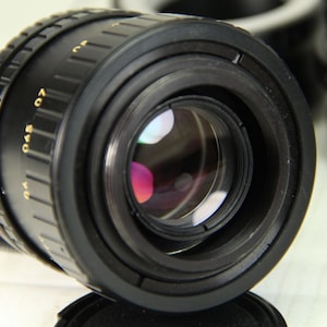 Puede incluir: Un objetivo de c&aacute;mara negro con marcas doradas, que presenta un reflejo multicolor en el cristal. El objetivo tiene un anillo de enfoque estriado y est&aacute; inclinado hacia el espectador. Otros accesorios de c&aacute;mara son visibles en el fondo.