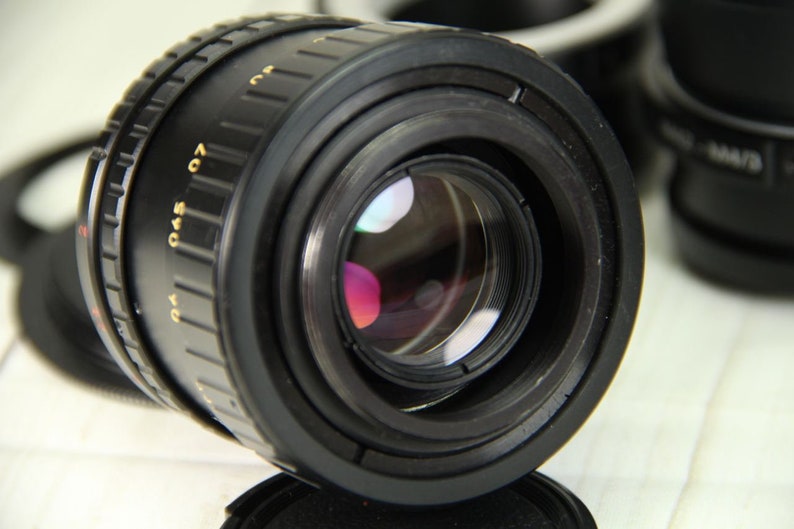 Puede incluir: Un objetivo de c&aacute;mara negro con marcas doradas, que presenta un reflejo multicolor en el cristal. El objetivo tiene un anillo de enfoque estriado y est&aacute; inclinado hacia el espectador. Otros accesorios de c&aacute;mara son visibles en el fondo.