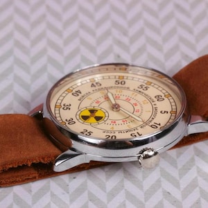May include: A silver wristwatch with a brown leather band. The watch face is white with black numbers and a yellow and black symbol in the center. The text on the watch face reads "СССР ЗАВОД ИМ. ПЕРВОГО МАЯ".