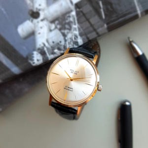 K&ouml;nnte beinhalten: Eine goldfarbene Armbanduhr mit wei&szlig;em Zifferblatt und schwarzem Lederarmband. Das Zifferblatt der Uhr hat schwarze r&ouml;mische Ziffern als Stundenmarkierungen, schwarze Zeiger und den Text "Poljot de Luxe" und "23 jewels shockproof".