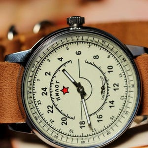 Puede incluir: Un reloj de pulsera plateado con una correa de cuero marr&oacute;n. La esfera del reloj es blanca con marcas negras y la palabra "Пилот" en escritura cir&iacute;lica. Una estrella roja se encuentra en el centro de la esfera.