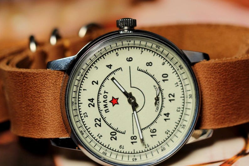 Puede incluir: Un reloj de pulsera plateado con una correa de cuero marr&oacute;n. La esfera del reloj es blanca con marcas negras y la palabra "Пилот" en escritura cir&iacute;lica. Una estrella roja se encuentra en el centro de la esfera.