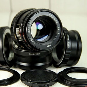 Puede incluir: Colecci&oacute;n de lentes y adaptadores de c&aacute;mara negros, incluido un lente marcado "MC ГЕЛИОС 44-3 2/58". Varios adaptadores est&aacute;n etiquetados como "M42-FX", "M42-NEX" y "M42-M4/3". Las lentes y adaptadores est&aacute;n sobre una superficie de madera clara.