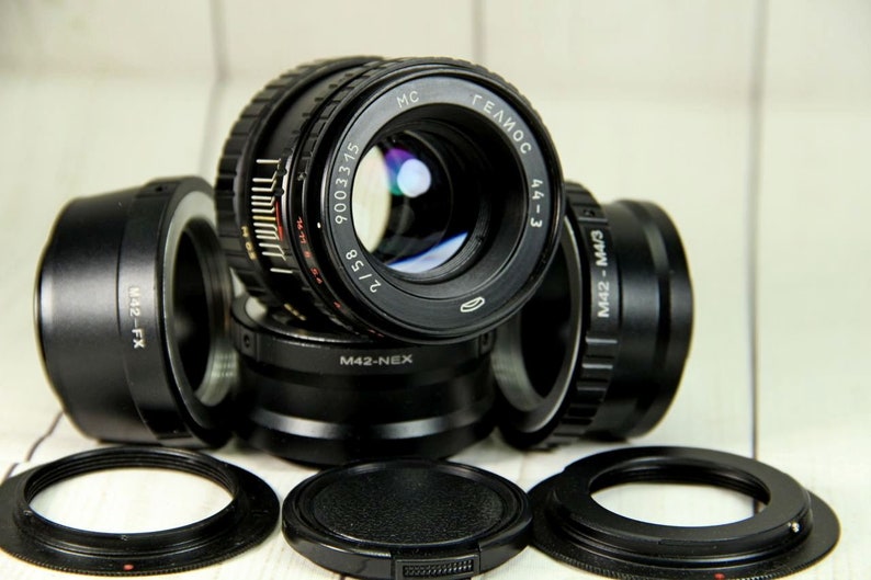 Puede incluir: Colecci&oacute;n de lentes y adaptadores de c&aacute;mara negros, incluido un lente marcado "MC ГЕЛИОС 44-3 2/58". Varios adaptadores est&aacute;n etiquetados como "M42-FX", "M42-NEX" y "M42-M4/3". Las lentes y adaptadores est&aacute;n sobre una superficie de madera clara.